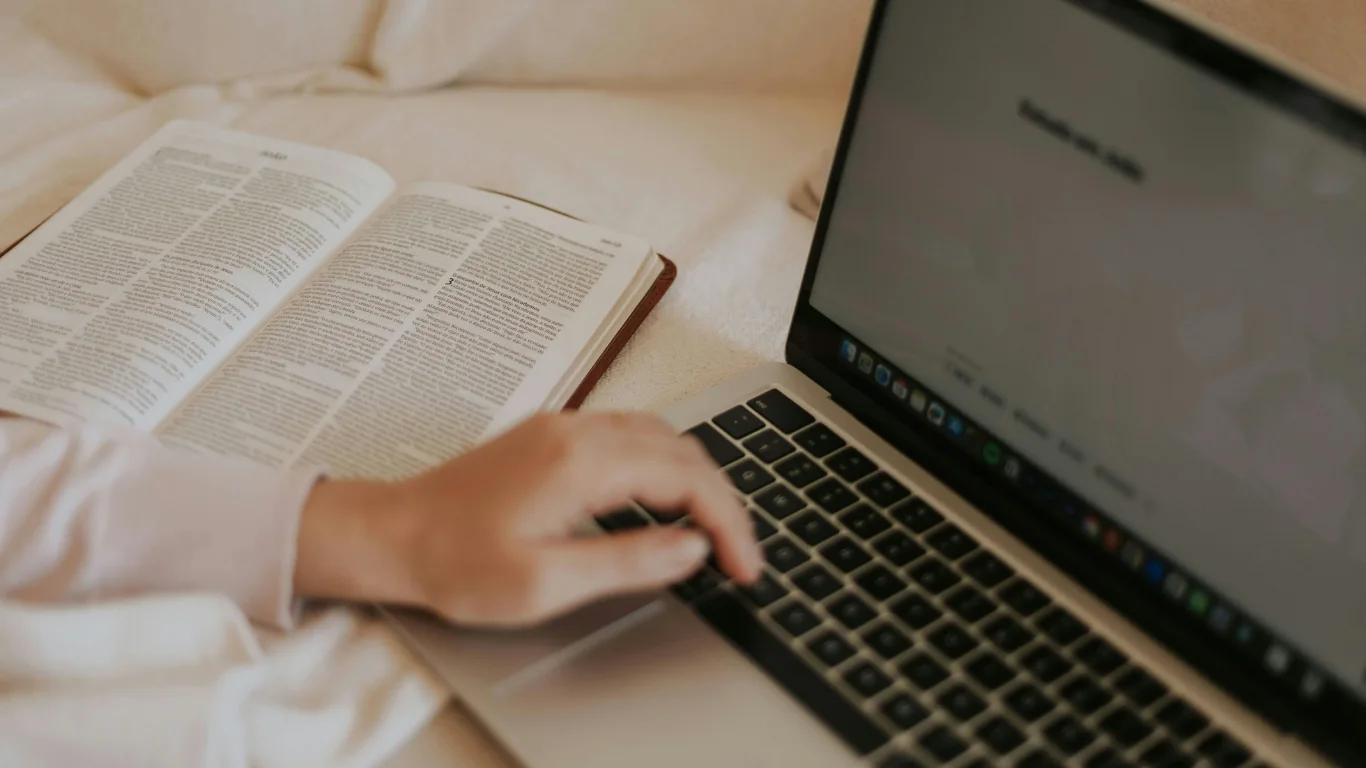Main sur le clavier d'un ordinateur portable près d'un livre ouvert, sur un plaid crème
